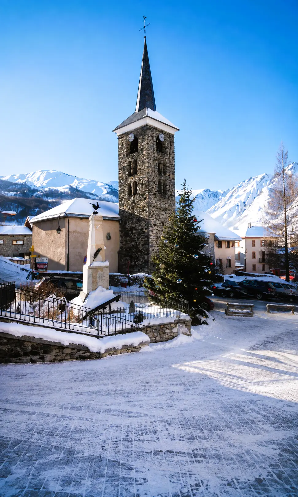 Église de Saint-Martin-de-Belleville sous la neige, avec son clocher en pierre, le monument aux morts et les montagnes de Savoie en arrière-plan.